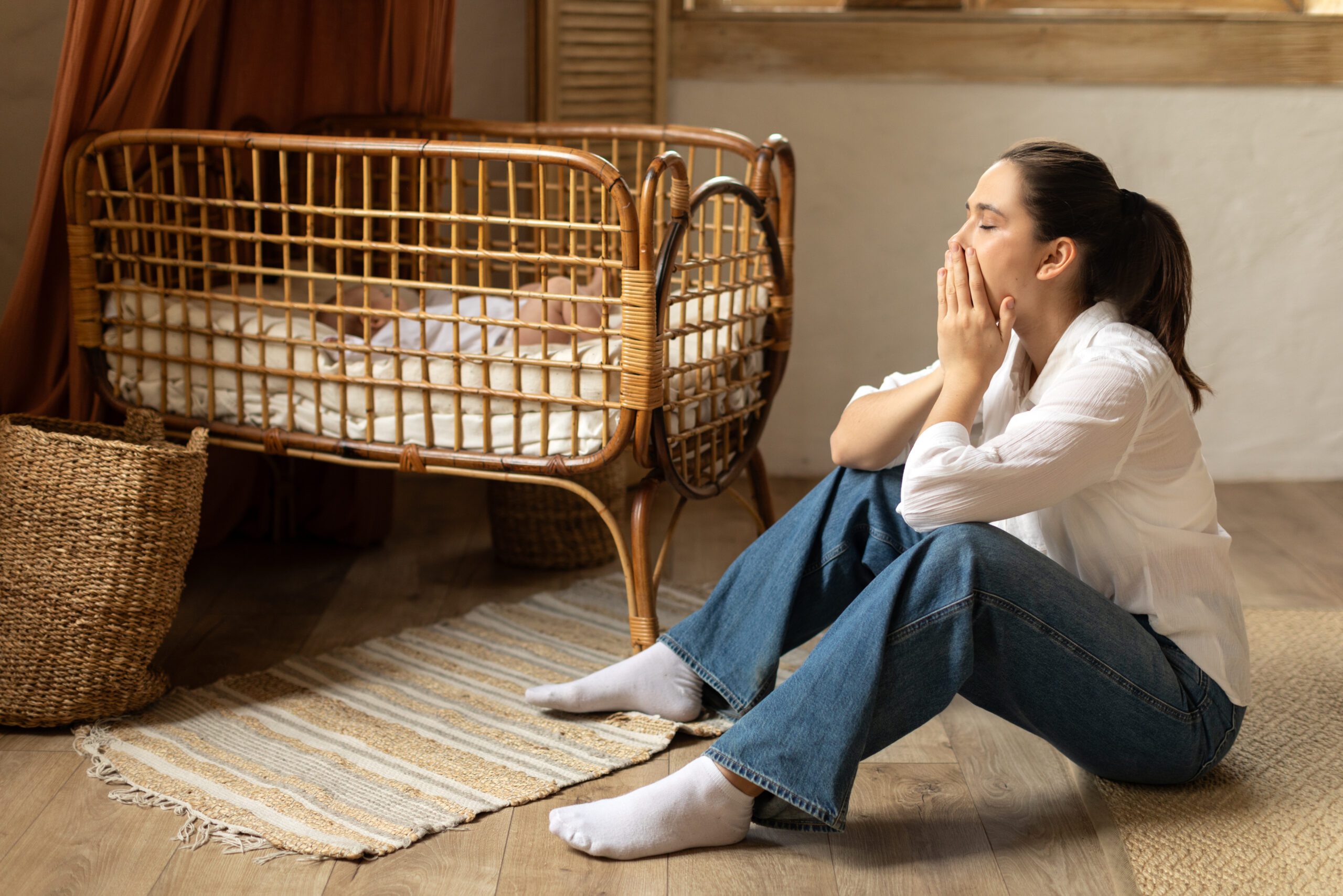 Young caucasian tired woman sitting on the floor near children’s cot, baby sleeping in her cradle. Postnatal depression and stressful motherhood concept